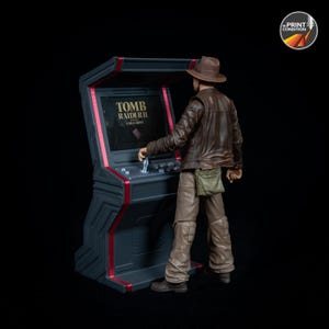 May include: A figurine of a person in a brown leather jacket and hat playing a Tomb Raider II arcade game. The arcade machine is black with red trim. The game screen displays the game's title.