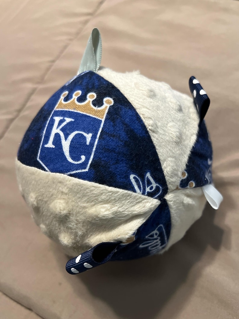 Puede incluir: Una pelota de tela azul y blanca con un centro blanco y mullido. La pelota tiene un patr&oacute;n azul y blanco con una corona dorada y las letras "KC" en un lado. La pelota tiene cuatro lazos azules y blancos con lunares.