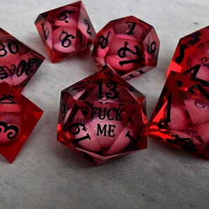 Peut inclure: Un ensemble de sept d&eacute;s poly&eacute;driques rouge translucide avec des chiffres noirs. Le d&eacute; &agrave; vingt faces porte l'inscription FUCK ME en lettres noires. Chaque d&eacute; contient un c&oelig;ur rose paillet&eacute; visible &agrave; travers la r&eacute;sine rouge transparente.
