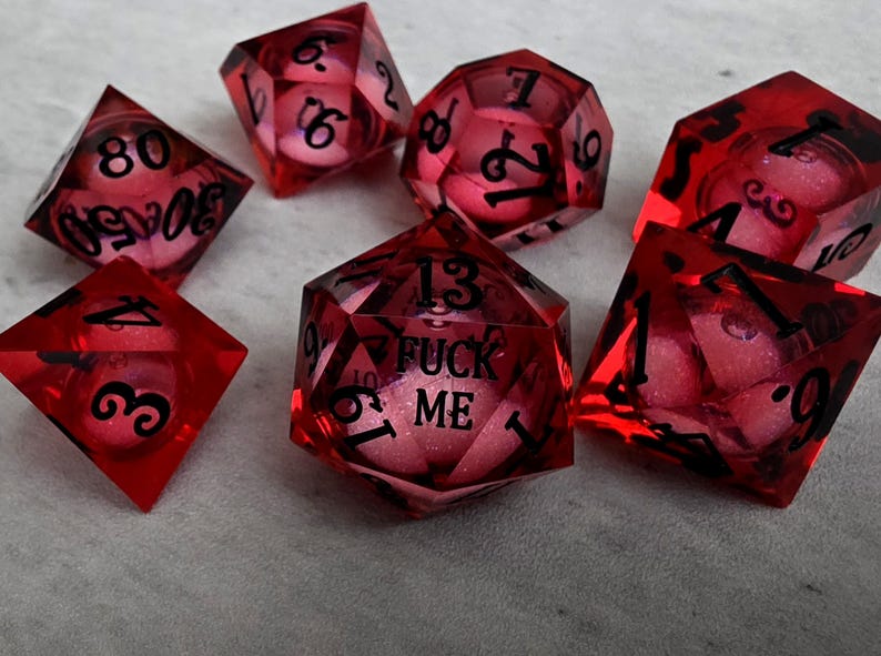 Peut inclure: Un ensemble de sept d&eacute;s poly&eacute;driques rouge translucide avec des chiffres noirs. Le d&eacute; &agrave; vingt faces porte l'inscription FUCK ME en lettres noires. Chaque d&eacute; contient un c&oelig;ur rose paillet&eacute; visible &agrave; travers la r&eacute;sine rouge transparente.