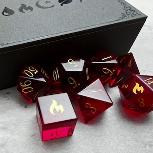 May include: A set of red, translucent polyhedral dice with gold numbering and symbols, including a flame icon on a six-sided die. The dice are arranged near a black box with decorative edges, suitable for tabletop gaming.
