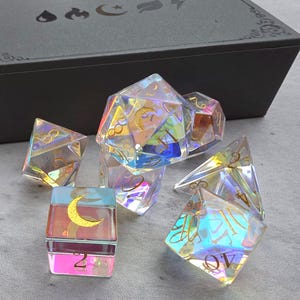 May include: A set of iridescent polyhedral dice with gold numbering and moon symbols, alongside a black box with decorative symbols. The dice are in various geometric shapes, reflecting light with a rainbow effect. The set includes a d20, d12, d10, d8, d6, and d4.