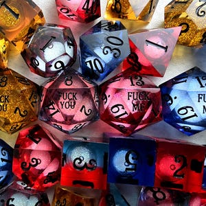 Peut inclure: Une collection de d&eacute;s poly&eacute;driques multicolores en r&eacute;sine translucide avec des paillettes internes. Plusieurs d&eacute;s pr&eacute;sentent des textes grav&eacute;s en noir, notamment les phrases FUCK YOU et FUCK ME. L'ensemble inclut diverses formes comme des d20 et d6.