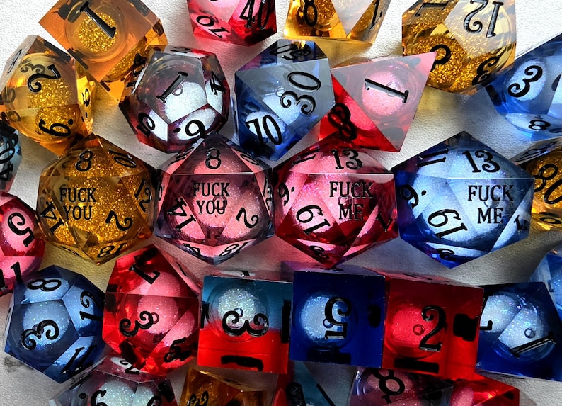 Peut inclure: Une collection de d&eacute;s poly&eacute;driques multicolores en r&eacute;sine translucide avec des paillettes internes. Plusieurs d&eacute;s pr&eacute;sentent des textes grav&eacute;s en noir, notamment les phrases FUCK YOU et FUCK ME. L'ensemble inclut diverses formes comme des d20 et d6.