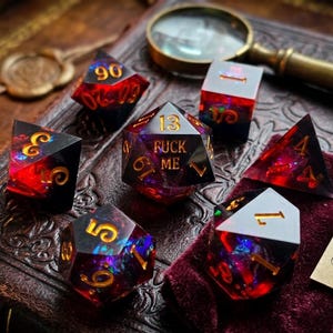 May include: A set of seven polyhedral dice featuring a dark, translucent resin base with vibrant red and blue internal inclusions and gold-colored numerals. The central twenty-sided die displays the text FUCK ME in place of the number twenty.