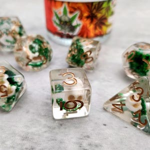 May include: A set of clear resin dice with green and white inclusions and copper-colored numbers. The dice are arranged on a light gray surface. A blurred image of a container with a cannabis leaf design is in the background.