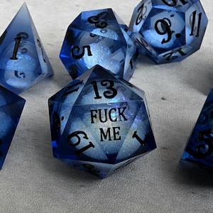 Peut inclure: Un ensemble de d&eacute;s poly&eacute;driques bleu translucide avec des chiffres noirs. Le d&eacute; &agrave; vingt faces central porte l'inscription FUCK ME en lettres noires. Chaque d&eacute; contient un c&oelig;ur de paillettes blanches visible &agrave; travers la r&eacute;sine bleue.