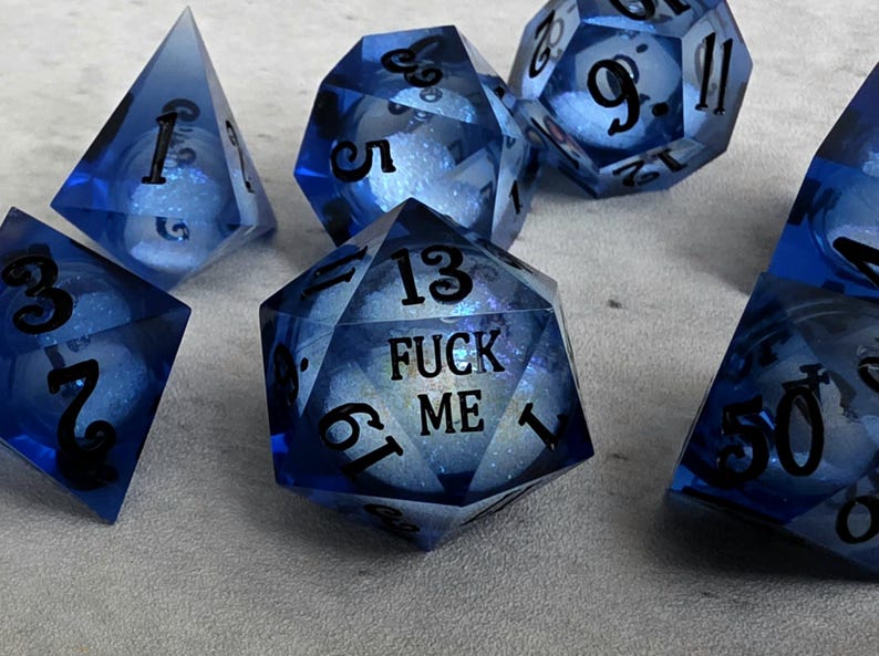 Peut inclure: Un ensemble de d&eacute;s poly&eacute;driques bleu translucide avec des chiffres noirs. Le d&eacute; &agrave; vingt faces central porte l'inscription FUCK ME en lettres noires. Chaque d&eacute; contient un c&oelig;ur de paillettes blanches visible &agrave; travers la r&eacute;sine bleue.