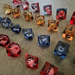 May include: A collection of multi-sided polyhedral dice in various translucent colours including red, blue, orange, and black. Each die features clear, dark-coloured numbers etched into the resin surfaces, showcasing a variety of geometric shapes.