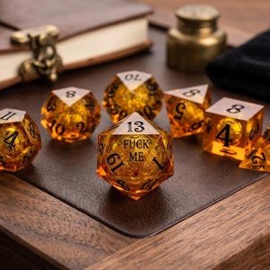 May include: A set of seven translucent amber-colored polyhedral dice with black numbering. The central twenty-sided die features the text FUCK ME printed in black ink. Each die contains a small, spherical gold-colored core suspended inside.