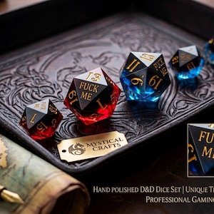 May include: A set of multi-faceted polyhedral dice featuring a black and translucent red or blue color scheme. Each die face displays gold-colored numbers and the text FUCK ME. A small gold-colored metal tag with the text MYSTICAL CRAFTS is included.