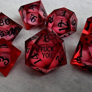 Peut inclure: Un ensemble de sept d&eacute;s poly&eacute;driques rouge translucide avec des chiffres noirs. Le d&eacute; &agrave; vingt faces porte l'inscription FUCK YOU &agrave; la place du chiffre vingt. Chaque d&eacute; contient une petite sph&egrave;re rose-rouge suspendue dans la r&eacute;sine rouge.