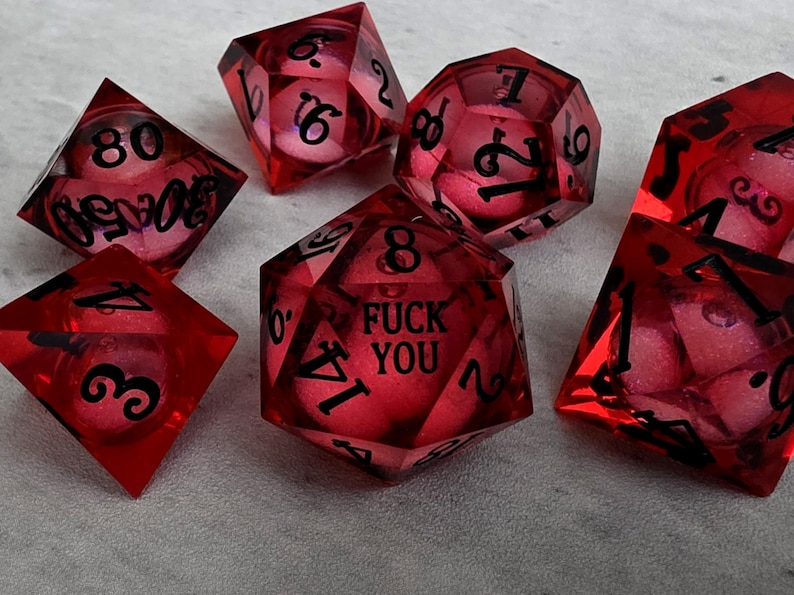Peut inclure: Un ensemble de sept d&eacute;s poly&eacute;driques rouge translucide avec des chiffres noirs. Le d&eacute; &agrave; vingt faces porte l'inscription FUCK YOU &agrave; la place du chiffre vingt. Chaque d&eacute; contient une petite sph&egrave;re rose-rouge suspendue dans la r&eacute;sine rouge.