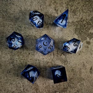 May include: A set of seven polyhedral dice featuring a translucent blue and white swirled resin design. The dice include black numbers and a crescent moon symbol on the twenty-sided die. The set consists of a d20, d12, d10, d8, d6, d4, and a percentile die.