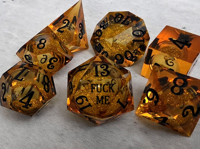 Peut inclure: Un ensemble de sept d&eacute;s poly&eacute;driques ambr&eacute;s translucides contenant des paillettes dor&eacute;es en suspension. Chaque d&eacute; pr&eacute;sente des chiffres grav&eacute;s en noir. Un d&eacute; &agrave; vingt faces affiche le texte FUCK ME en lettres noires sur sa face sup&eacute;rieure.