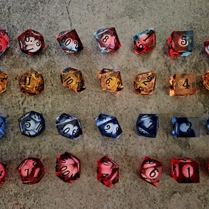 May include: A collection of twenty-eight polyhedral dice arranged in four rows of seven. The dice feature various translucent colours including red, orange, and blue, with internal spherical shapes and black numerical markings on their surfaces.