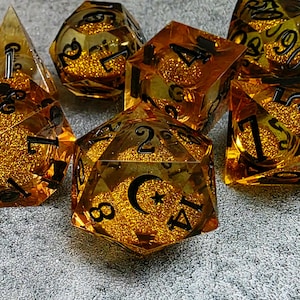 May include: Set of seven amber-colored polyhedral dice with black numbering and glitter inclusions. The dice are various shapes, including a d4, d6, d8, d10, d12, and d20. The d20 features a crescent moon and star symbol.