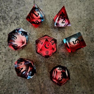 May include: A set of seven polyhedral dice featuring a translucent red resin base with swirling black and white smoke-like inclusions. Each die has black engraved numbers, and the twenty-sided die features a crescent moon symbol in place of the number twenty.