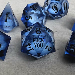 Peut inclure: Un ensemble de d&eacute;s poly&eacute;driques en r&eacute;sine bleue translucide avec des chiffres noirs. Chaque d&eacute; contient une sph&egrave;re blanche scintillante &agrave; l'int&eacute;rieur. Le d&eacute; &agrave; vingt faces au centre porte le texte FUCK YOU imprim&eacute; en noir au lieu du chiffre vingt.