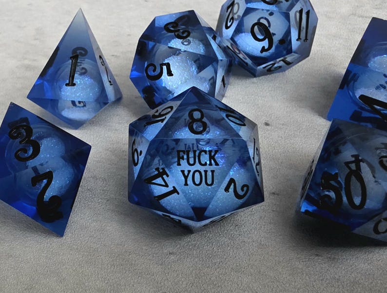 Peut inclure: Un ensemble de d&eacute;s poly&eacute;driques en r&eacute;sine bleue translucide avec des chiffres noirs. Chaque d&eacute; contient une sph&egrave;re blanche scintillante &agrave; l'int&eacute;rieur. Le d&eacute; &agrave; vingt faces au centre porte le texte FUCK YOU imprim&eacute; en noir au lieu du chiffre vingt.