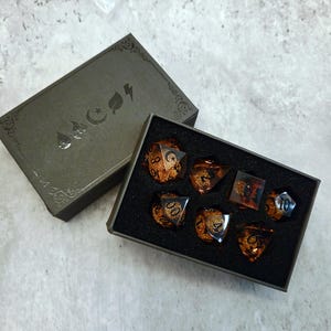 May include: A set of seven polyhedral dice with a translucent amber and gold glitter finish, featuring black numbers. The dice are housed in a black rectangular box with foam inserts, accompanied by a matching lid displaying four silver icons.
