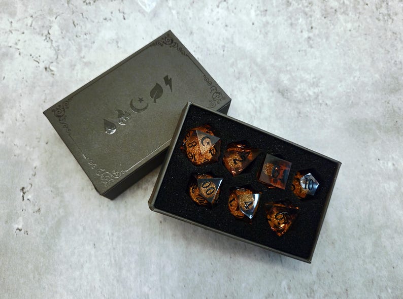 May include: A set of seven polyhedral dice with a translucent amber and gold glitter finish, featuring black numbers. The dice are housed in a black rectangular box with foam inserts, accompanied by a matching lid displaying four silver icons.