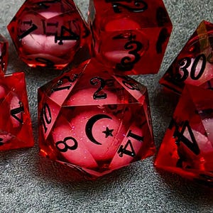 以下が含まれることがあります： 黒い数字と三日月と星のデザインが施された、赤色の多面体サイコロのコレクション。 サイコロは半透明の素材でできており、内部を垣間見ることができます。 サイコロはテーブルゲーム用です。