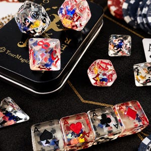 May include: A set of clear resin polyhedral dice containing colorful confetti and playing card suit symbols. The dice feature red and black numbers, with some pieces shaped like square tiles displaying spade, heart, club, and diamond icons.