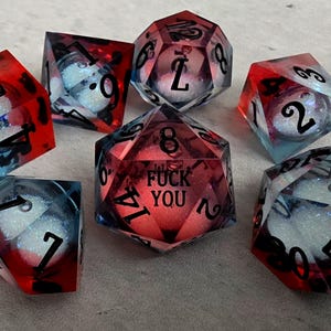 Peut inclure: Un ensemble de sept d&eacute;s poly&eacute;driques en r&eacute;sine translucide rouge et bleue. Chaque d&eacute; contient une sph&egrave;re blanche scintillante &agrave; l'int&eacute;rieur. Le d&eacute; &agrave; vingt faces au centre porte le texte noir FUCK YOU au lieu du chiffre vingt.