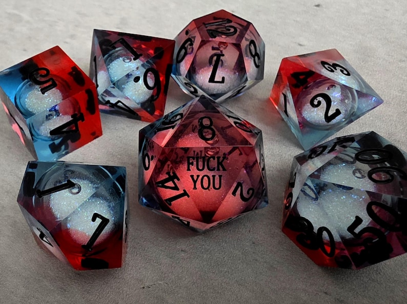 Peut inclure: Un ensemble de sept d&eacute;s poly&eacute;driques en r&eacute;sine translucide rouge et bleue. Chaque d&eacute; contient une sph&egrave;re blanche scintillante &agrave; l'int&eacute;rieur. Le d&eacute; &agrave; vingt faces au centre porte le texte noir FUCK YOU au lieu du chiffre vingt.