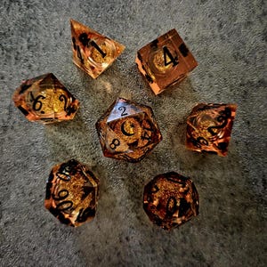 May include: A set of seven polyhedral dice made of translucent amber-coloured resin with embedded gold-coloured glitter flakes. Each die features sharp edges and clear black-inked numbers for readability, including a d4, d6, d8, d10, d12, d20, and percentile die.