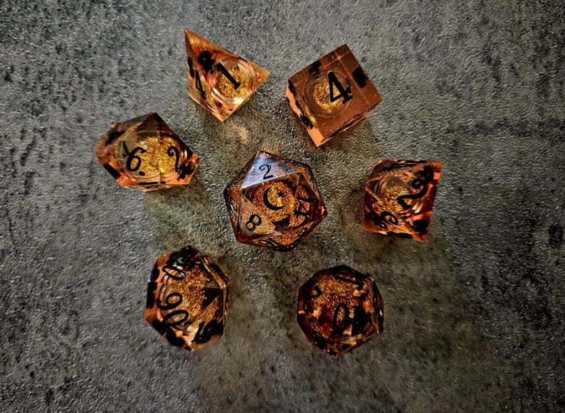 May include: A set of seven polyhedral dice made of translucent amber-coloured resin with embedded gold-coloured glitter flakes. Each die features sharp edges and clear black-inked numbers for readability, including a d4, d6, d8, d10, d12, d20, and percentile die.
