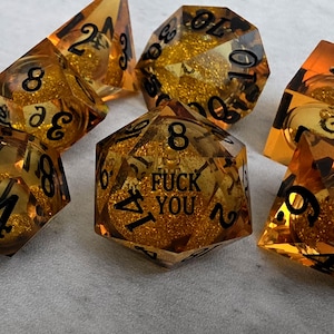 Peut inclure: Un ensemble de sept d&eacute;s poly&eacute;driques ambr&eacute;s translucides contenant des paillettes dor&eacute;es en suspension. Chaque d&eacute; pr&eacute;sente des chiffres grav&eacute;s en noir. Le d&eacute; &agrave; vingt faces au centre affiche le texte FUCK YOU &agrave; la place du chiffre vingt.