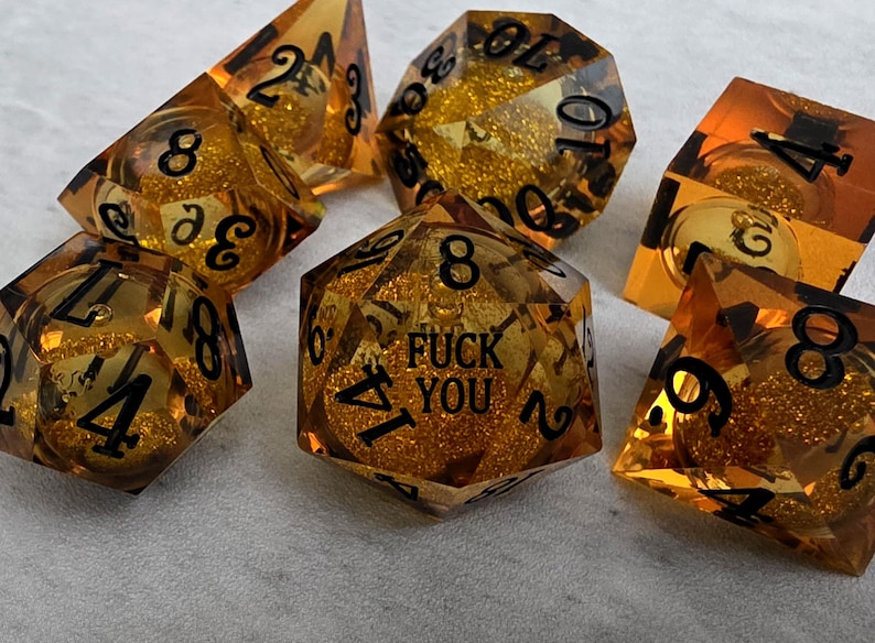 Peut inclure: Un ensemble de sept d&eacute;s poly&eacute;driques ambr&eacute;s translucides contenant des paillettes dor&eacute;es en suspension. Chaque d&eacute; pr&eacute;sente des chiffres grav&eacute;s en noir. Le d&eacute; &agrave; vingt faces au centre affiche le texte FUCK YOU &agrave; la place du chiffre vingt.