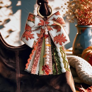 May include: A decorative bag charm featuring a large bow with a white and orange autumn leaf pattern, attached to a gold-toned metal ring. It includes various hanging ribbons in orange, green, and brown, along with a strand of white beads.