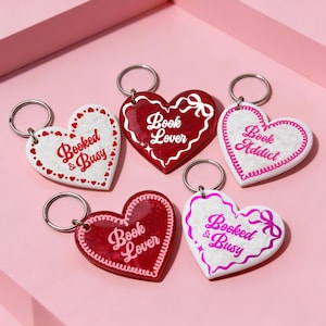 May include: Heart-shaped keychains in red and white with phrases like "Book Lover" and "Book Addict." Each keychain has a silver keyring. The keychains are on a pink surface.