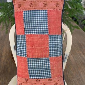 May include: A rectangular quilted pillow featuring a patchwork design of coral-colored fabric with small brown spiral patterns and blue and white gingham check squares. The pillow is finished with a dark blue piped edge and visible stitching.