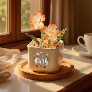 May include: A white ceramic planter with a floral and book design, holding artificial flowers with orange and white petals. The planter sits on a wooden tray, with the text "Mrs. Smith" visible. A cup and saucer are in the background.