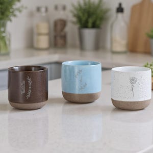 Pode incluir: Três canecas de cerâmica num ambiente de cozinha. Uma é castanha com o nome "Natalia" e um desenho de folhas, outra é azul clara com um desenho floral, e a terceira é branca com um desenho floral. Todas têm uma base castanha.
