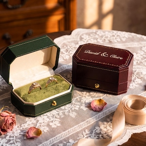 May include: Two octagonal jewelry boxes, one dark green and one burgundy, featuring gold-tone latches. The open green box contains two gold rings on a green velvet insert. The closed burgundy box is engraved with the text David & Claire.