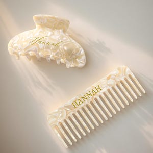 Op de afbeelding: Een crèmekleurige haarclip en kam set. Beide items hebben een gemarmerd patroon en de naam "HANNAH" in gouden letters. De haarclip is ovaal van vorm en de kam heeft brede tanden.
