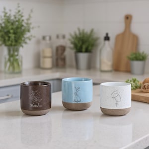 May include: Three ceramic mugs with a speckled, two-tone design. One is brown with a moon and floral design and the name "Keisha". Another is light blue with a floral design and the name "Sofia". The third is white with a floral design and the name "Sofia".