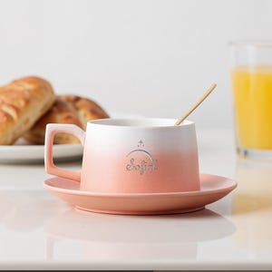 May include: A ceramic coffee cup and saucer set with a gradient color scheme, transitioning from white to peach. The cup has a handle and the word "Sofia!" is printed on the front. A small wooden spoon rests inside the cup. The set is on a white surface.