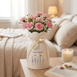 May include: A cream-colored vase with a scalloped top, tied with a gold ribbon, holds pink carnations and white daisies. The vase is inscribed with "Mama's Garden" and names. A lit candle and open book are nearby.