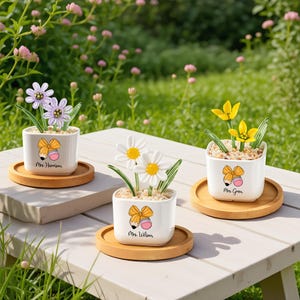 May include: Three white ceramic flower pots with decorative flowers, each on a round wooden coaster. The pots have a pencil and bow design with names printed below. The flowers are purple, white, and yellow.