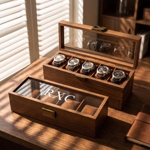 May include: Two rectangular wooden watch display cases with glass lids. The front case features the white engraved letters RXC. The back case holds five wristwatches on brown cushions and displays an engraved laurel wreath with the letter A.