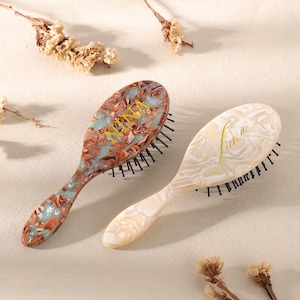 May include: Two oval-shaped hairbrushes. One brush has a brown, blue, and white marbled design, and the other is off-white with a marbled pattern. Both brushes have black bristles and the word "Juha" in a yellow script.