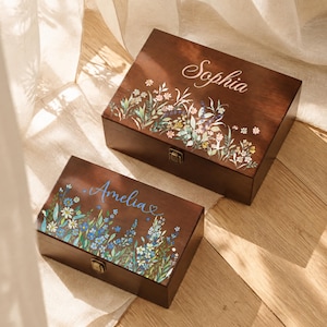 May include: Two rectangular dark brown wooden keepsake boxes with metal latches. The larger box features the name Sophia in white script and a colorful wildflower print. The smaller box features the name Amelia in blue script and a blue floral print.