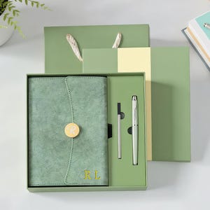 Puede incluir: Un juego de regalo verde salvia que contiene un diario, un bolígrafo y accesorios. El diario tiene una cubierta texturizada con un botón dorado y las iniciales "RL". El bolígrafo es plateado. El juego se presenta en una caja verde con una bolsa de regalo a juego.