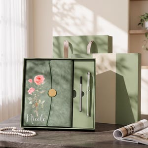 May include: A sage green journal set featuring a floral rose illustration and the name Nicole printed in white script. The set includes a matching green pen and a slim black stylus, all housed in a coordinated green gift box with a ribbon handle.